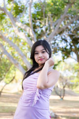 Half body portrait of an asian woman wearing a purple shirt, smiling in front of trees at a park near a beach in Nusa Dua, Bali.