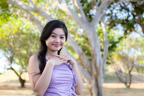 Half body portrait of an asian woman wearing a purple shirt, smiling in front of trees at a park near a beach in Nusa Dua, Bali.