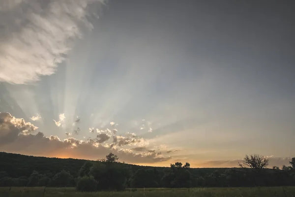 The sun\'s rays of the evening setting sun break through the clouds from behind the forest hill