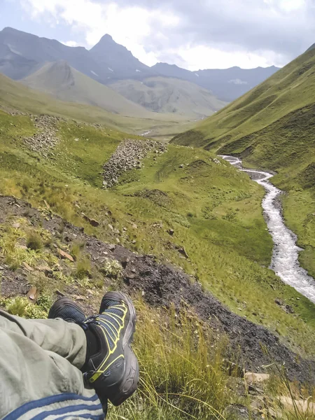 A stormy mountain river flows between green hilly meadows and ridges of the Caucasian mountain system against the backdrop of the pyramidal mountain Pabaku in cloudy weather