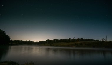 Gölün üzerinde yıldızlı bir gece ve sahildeki ağaçlar uzun pozlarda, bulutsuz bir yaz gecesinde