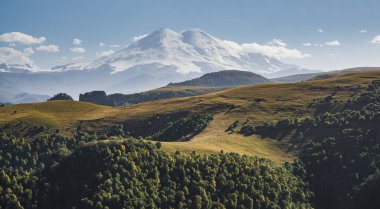 Kafkasya 'da sıcak bir sonbahar günü olan Elbrus Dağı' nın arka planına karşı parlak güneşin altında yaprak döken ağaçlardan ve dağ sıralarından oluşan bir ormanla dağ uçurumları.