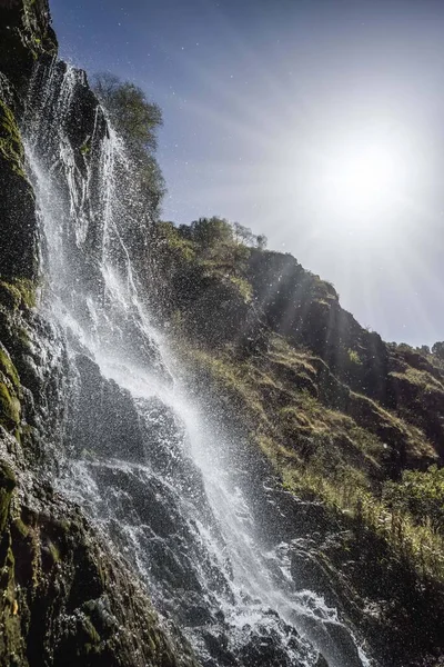 A mountain waterfall from a cold river flows over the rocks and splashes, illuminated by the bright sun, a cold waterfall between the hills of the Caucasus