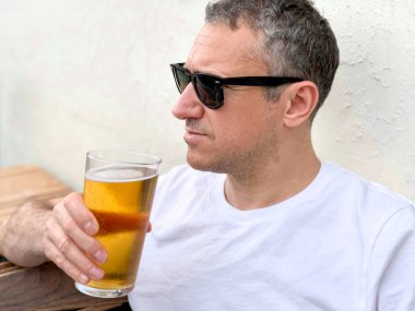 Orta yaşlı bir adam dışarıdaki bir kafe ya da restoran barında bira içiyor. Alkol içmek. Samimi. İçecek, yaz tatili, bar bahçesi. Beyaz tişört ve güneş gözlüğü takıyor. Bira ya da bira içmek.
