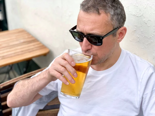 Middle aged man is drinking beer in a cafe or restaurant pub outdoor. Drinking alcohol. Candid. Refreshment, summer time, pub garden. wearing white t-shirt and sunglasses. Drinking lager or ale beer