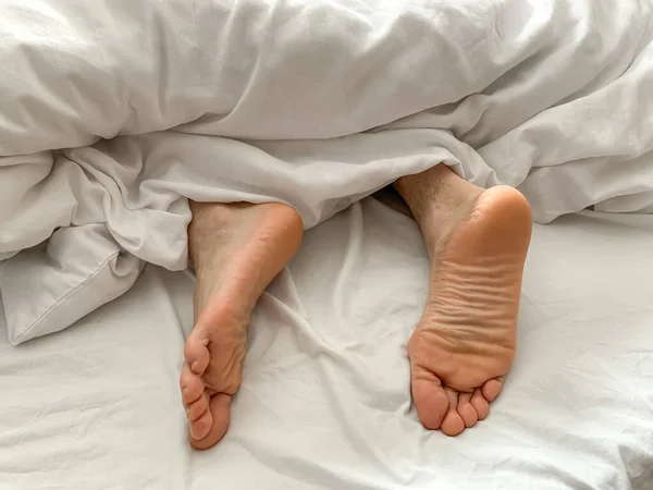 View of male feet, man sleeping at home. Middle aged adult man is sleeping at home. Cozy bedroom vibes. White bedsheets, bed linen, alone, early morning atmosphere.