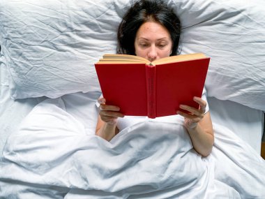 Young millennial candid woman is reading red book at home on white striped bedsheets. Early morning. Text in book is blurred. Education or slow living concept. Young adult beautiful woman