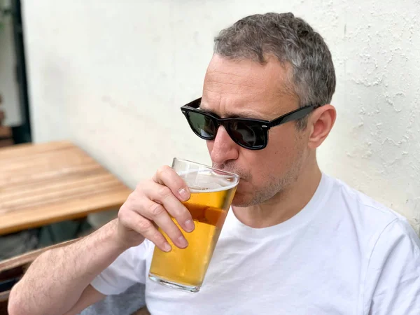 Middle aged man is drinking beer in a cafe or restaurant pub outdoor. Drinking alcohol. Candid. Refreshment, summer time, pub garden. wearing white t-shirt and sunglasses. Drinking lager or ale beer