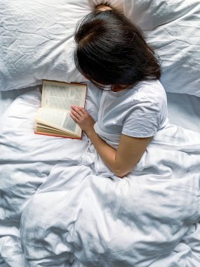 Young millennial candid woman is reading red book at home on white striped bedsheets. Early morning. Text in book is blurred. Education or slow living concept. Young adult beautiful woman
