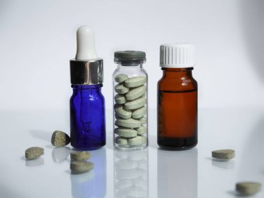 Yeşil ilaç haplı cam kavanoz, tıbbi damlalı mavi pipet ve beyaz ayna arka planında sıvı yağ bulunan kahverengi tıbbi şişe.