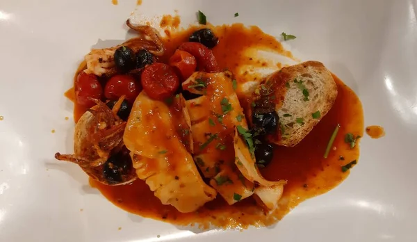 Domates soslu ızgara balık, zeytin, maydanoz ve beyaz seramik tabakta kızarmış ekmek.