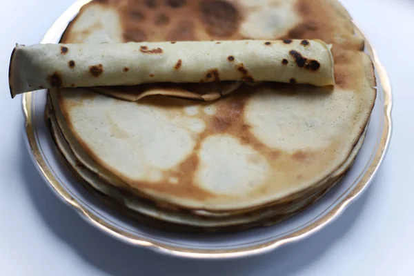a lot of ruddy pancakes and a pancake roll on top of them lie on a white plate with a golden border on a white kitchen table.for banners of cookbooks and notebooks
