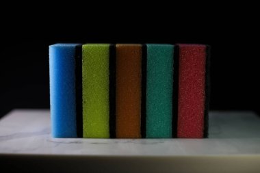 Tabakları yıkamak için mutfak süngerleri ve farklı renkte sarı mavi turuncu ve pembe yüzeyler koyu arkaplan üzerinde gri çizgiler bulunan beyaz mermer bir tahtada uzanıyor.