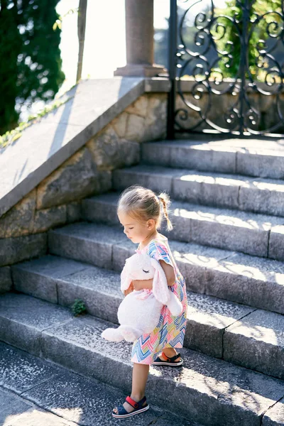 Little girl with a soft toy hare descends the stone steps in the garden. High quality photo