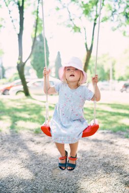 Little girl in a panama sits on a swing and holds on to the ropes with her hands. High quality photo