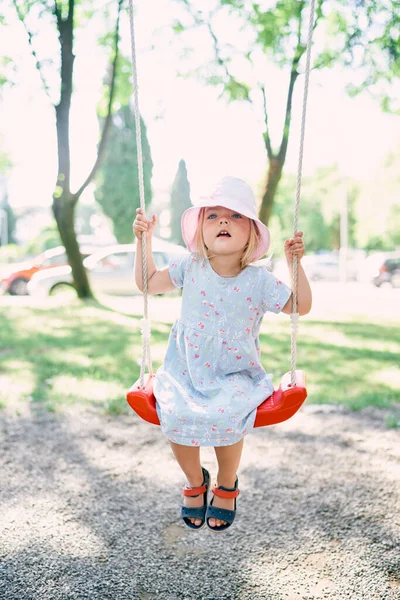 Little girl in a panama sits on a swing and holds on to the ropes with her hands. High quality photo