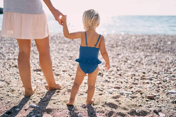 Little girl in a swimsuit holding her mom hand while standing on the beach. High quality photo