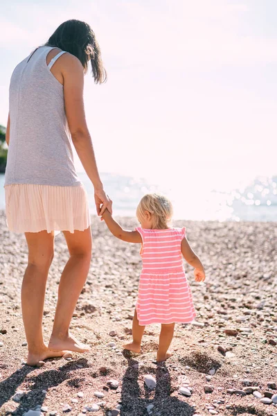 Mom and little girl walk barefoot along the beach holding hands. Back view. High quality photo