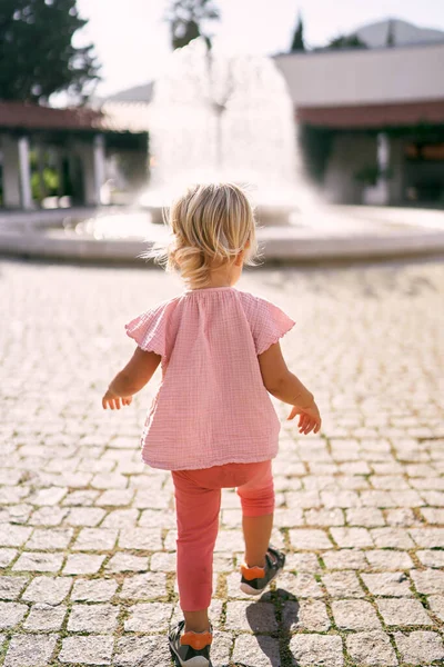 Little girl walks along the paving stones in the square towards the fountain. Back view. High quality photo