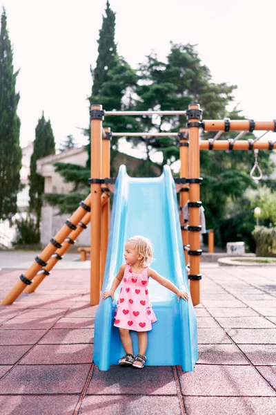 Little girl stands near the slide on the playground holding on to the sides. High quality photo