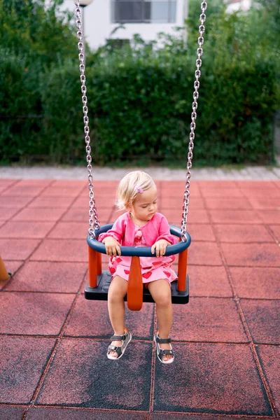 Little girl sits on a chain swing and holds on to a round handrail. High quality photo