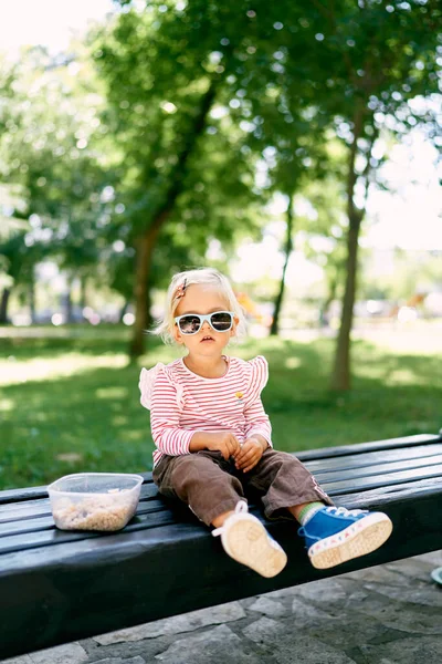 Little girl in sunglasses sits on a bench next to a lunch box. High quality photo