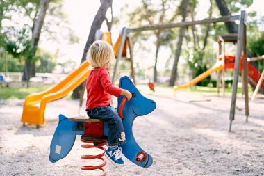 Little girl sits on a spring swing and looks at the playground. Back view. High quality photo