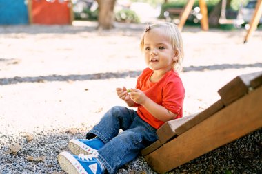 Little girl sits on the gravel near the wooden stairs on the playground. High quality photo