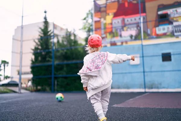 Little girl in a tracksuit is chasing the ball on the sports ground. High quality photo