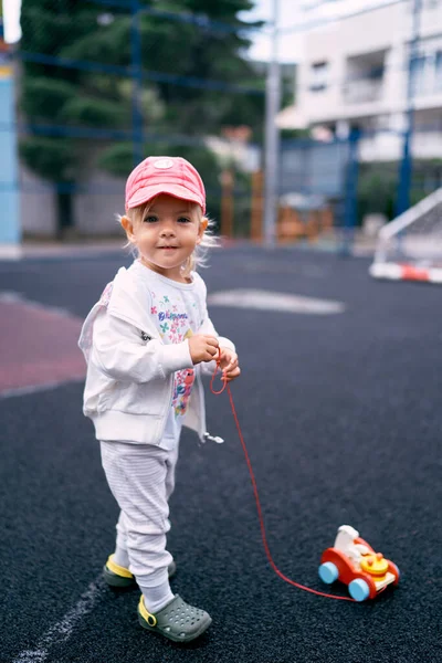 Little girl in a red cap stands on the sports ground with a toy car. High quality photo
