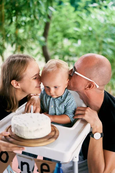 Mom and dad kiss a little boy on both sides sitting in front of a cake. High quality photo