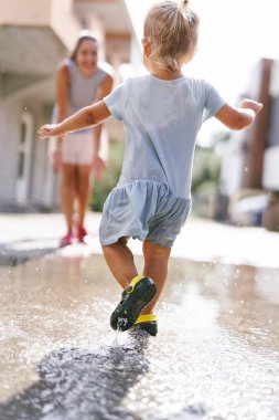 Küçük kız bir su birikintisinin içinden gülümseyen bir anneye koşuyor. Yüksek kalite fotoğraf