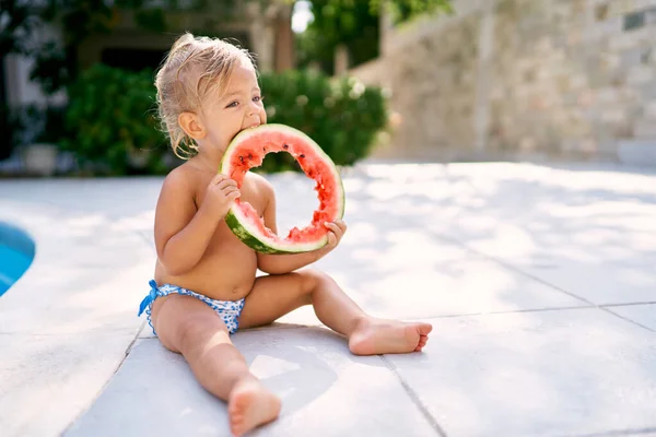 Küçük kız çitin yanındaki fayansta otururken yuvarlak bir karpuz parçasında delik açıyor. Yüksek kalite fotoğraf