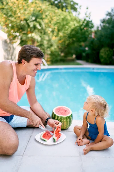 Babam havuz kenarında oturan küçük bir kızın önünde karpuzu dilimlere ayırır. Yüksek kalite fotoğraf