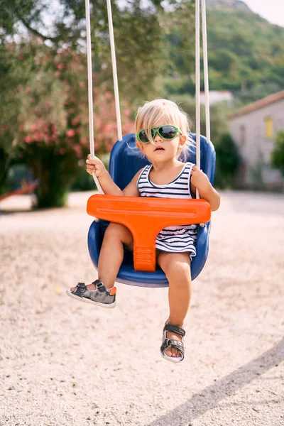Güneş gözlüklü küçük kız renkli bir salıncakta oturup iplere tutunuyor. Yüksek kalite fotoğraf