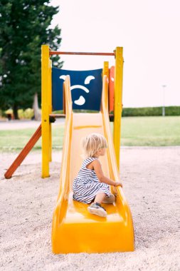 Küçük kız bir çocuğun kaydırağının dibinde yan oturup oyun parkına bakıyor. Yüksek kalite fotoğraf