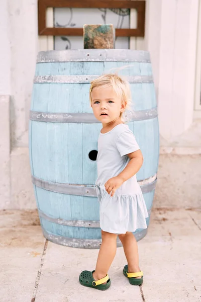 Küçük kız evin duvarına yakın mavi bir tahta varilin yanında duruyor. Yüksek kalite fotoğraf