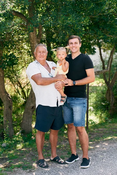 Babam ve büyükbabam küçük bir kızı yeşil bir ağacın gölgesinde kollarında tutuyorlar. Yüksek kalite fotoğraf