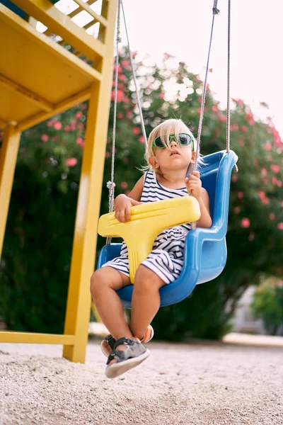 Küçük kız oyun parkında salıncakta bir gül ağacının arka planında oturuyor. Yüksek kalite fotoğraf