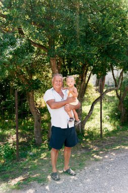 Büyükbaba, çitin arkasındaki yeşil ağacın yanında küçük bir kız tutuyor. Yüksek kalite fotoğraf