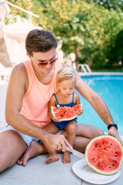 Babam havuz kenarında kucağında oturan küçük bir kızla karpuz kesiyor. Yüksek kalite fotoğraf