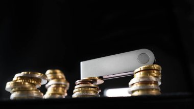 A crypto hardware wallet sitting on top of a stack of euro coins illuminated by a white light source that is visible in front of a black background