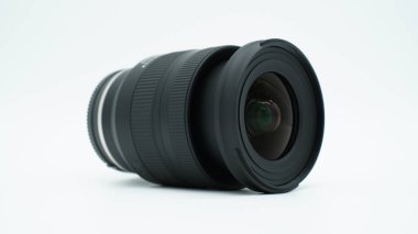 A black 11-20mm zoom lens for APSC cameras, lying on a white surface in front of a white background, with extended zoom, casting shadows on the surface.