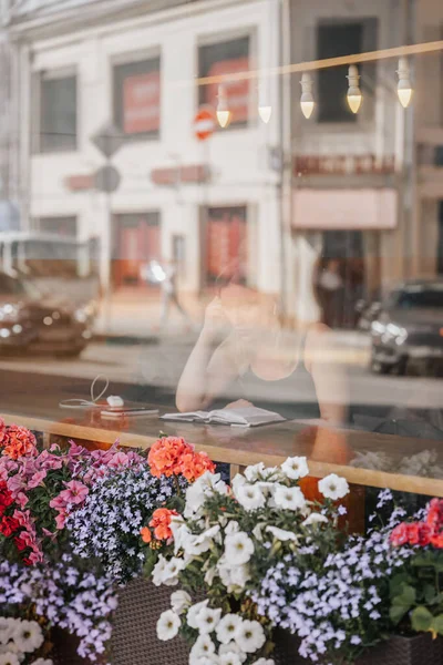 Detrás de la ventana. Chica en la mesa con libro, reflejo de la calle de la ciudad en el día de verano, borrosa bokeh — Foto de Stock