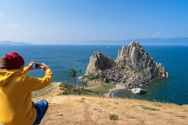 Unrecognizable girl back to us, sitting on high hill photographing rocky island mount.