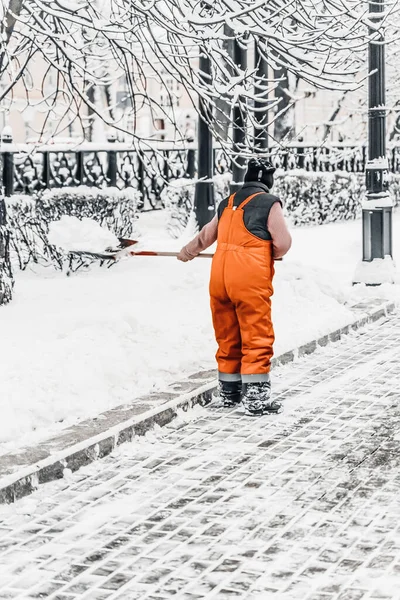 Operaio in uniforme arancione con pala pulizia strada. Rimozione della neve nella città invernale — Foto Stock