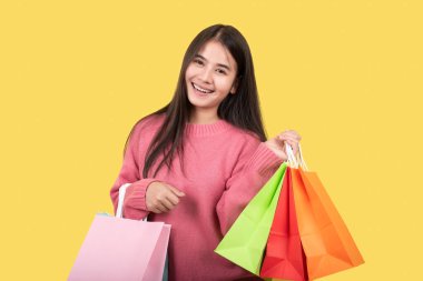 Çekici, gülümseyen, pembe tişörtlü, elinde alışveriş torbalarıyla sarı arka planda poz veren güzel bir kadın.