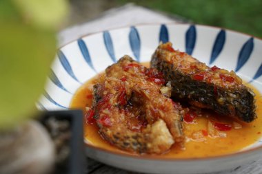 Eski ahşap bir zeminde seramik tabakta acılı kızarmış balık. Tadı ekşi, tatlı ve biraz baharatlı..