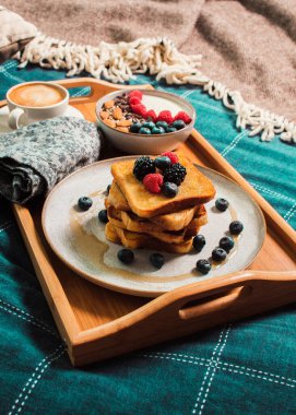 Cozy breakfast in bed, yogurt bowl with berries and nuts, french toasts, coffee cup on wooden plate. Close up still life. Food photography.