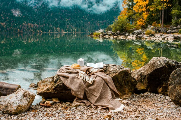 Picnic on stones in the Bavarian mountains, Germany. Plaid, cacao with marshmallow, croissant, book near lake and autumn forest. Cozy morning. Alps landscape. Breakfast in nature.